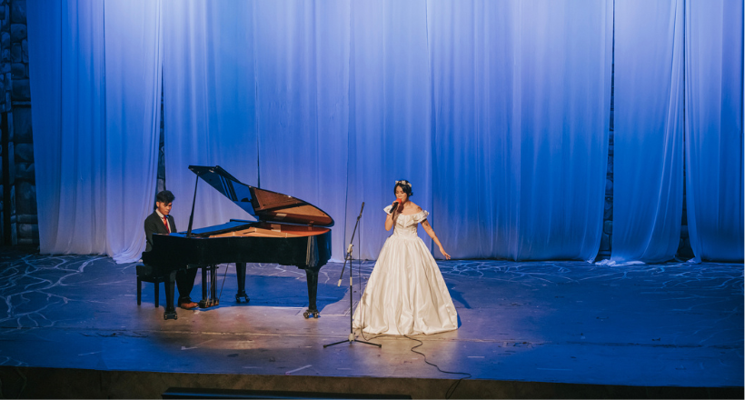A women sings and a man plays the piano during a live performance.
