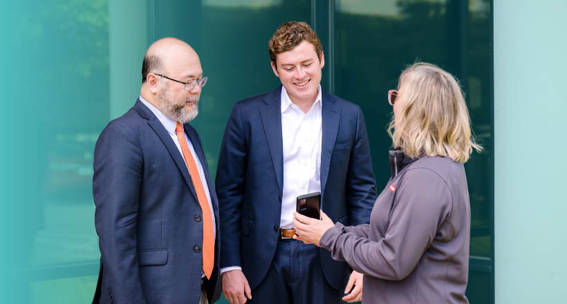 Howard, Chris, and Liz, three members of the intelligence and security team stand in the courtyard as they look a phone being held by Liz.