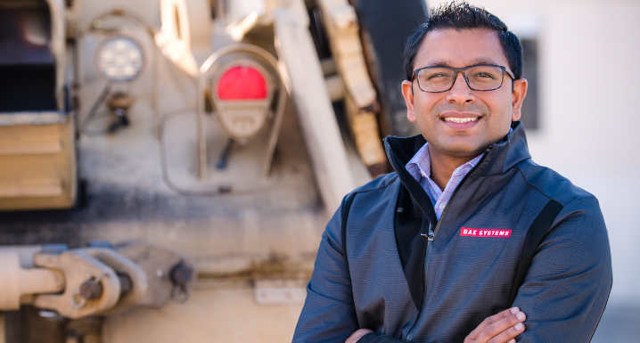 Harshit, an electrical engineer, stands in front of a combat vehicle as he poses for a photo. His arms are crossed and he's wearing a BAE Systems logoed jacket.