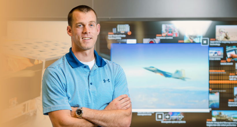 Nick, a systems engineer, smiles for a photo in front a screen showing a military jet.