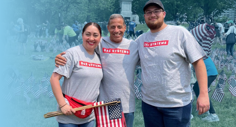 BAE Systems employees smile for a photo at a community investment event supporting our nation's troops. The employee on the left is holding three small American flags.
