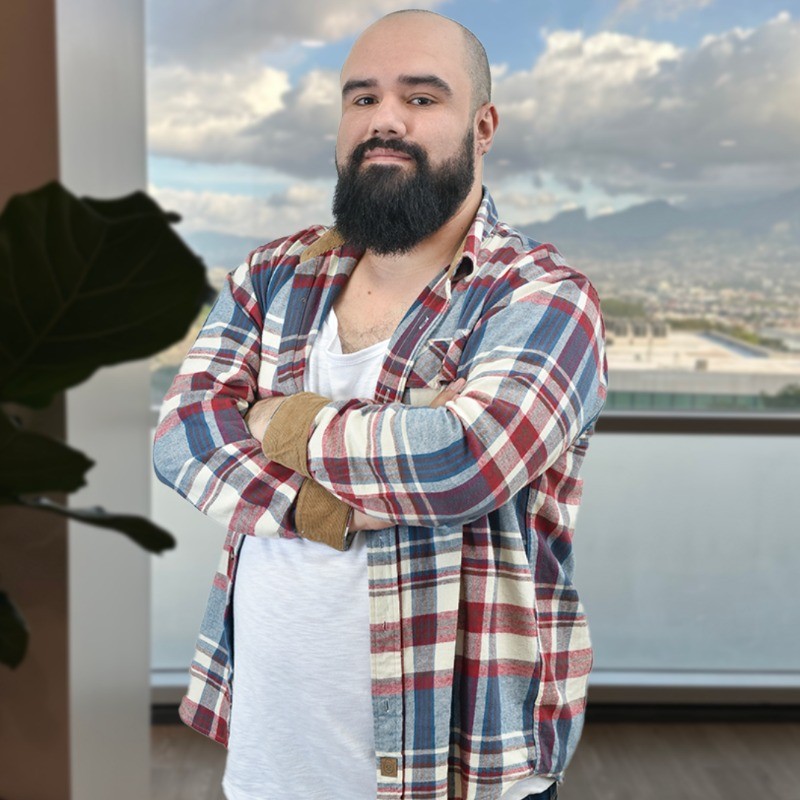 A man with a shaved head and a thick, dark beard stands confidently with his arms crossed. He is wearing a red, white, and blue plaid shirt over a white t-shirt. The background shows a large window with a view of a cityscape and mountains under a cloudy sky. A large green plant is partially visible in the foreground to the left.