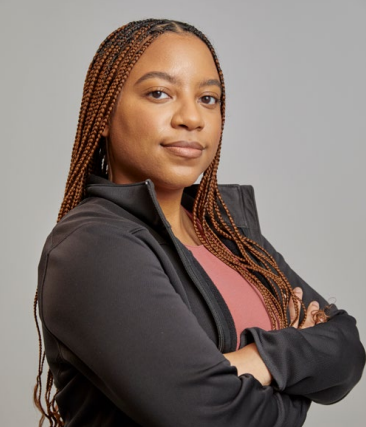 A woman with long, braided hair stands confidently with her arms crossed. She is wearing a black jacket over a pink top. She has a calm expression on her face and is looking directly at the camera. The background is a plain light gray color.