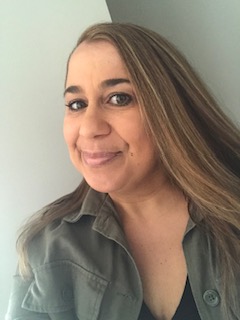 A woman with long, light brown hair is smiling slightly at the camera. She is wearing a green jacket over a black top. The background is plain and light-colored, with a slight shadow cast behind her.