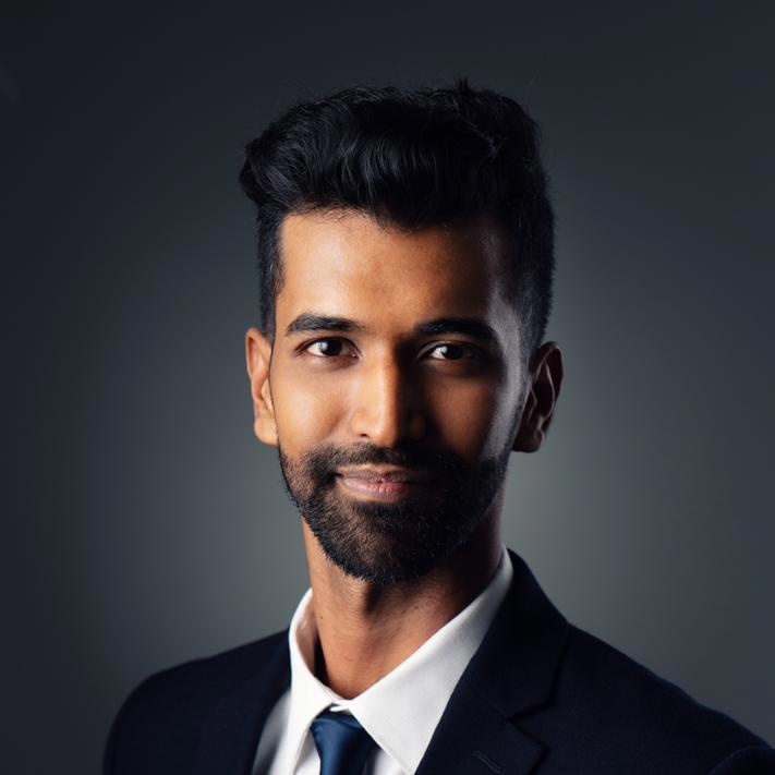 A close-up photo of a man with short, styled black hair and a neatly trimmed beard. He is wearing a dark suit, white shirt, and a dark tie. He is smiling subtly and looking slightly off-camera. The background is a dark gradient, which highlights his face.