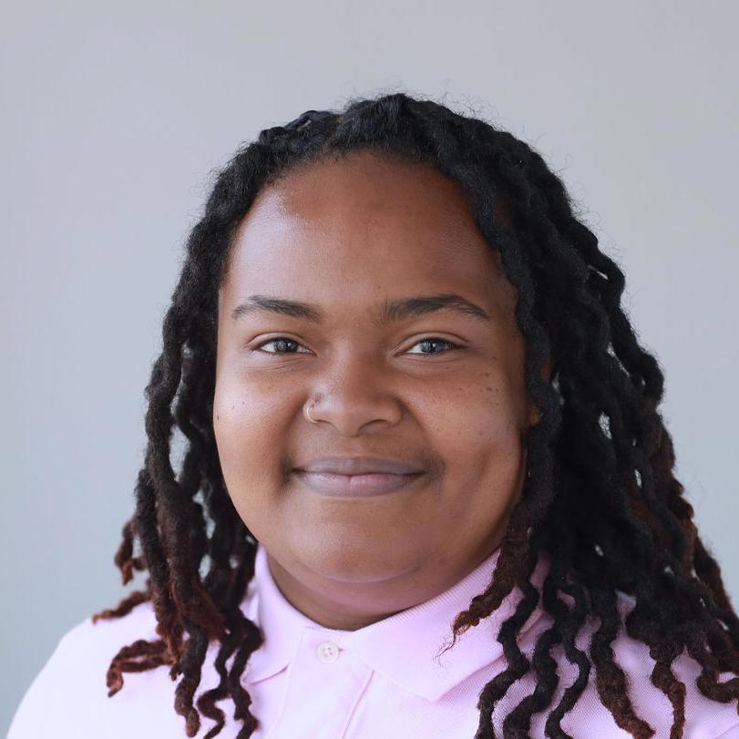 A close-up photo of a woman with long, dark dreadlocks and a nose piercing. They are smiling slightly and wearing a light pink collared shirt. The background is a plain, light gray color.