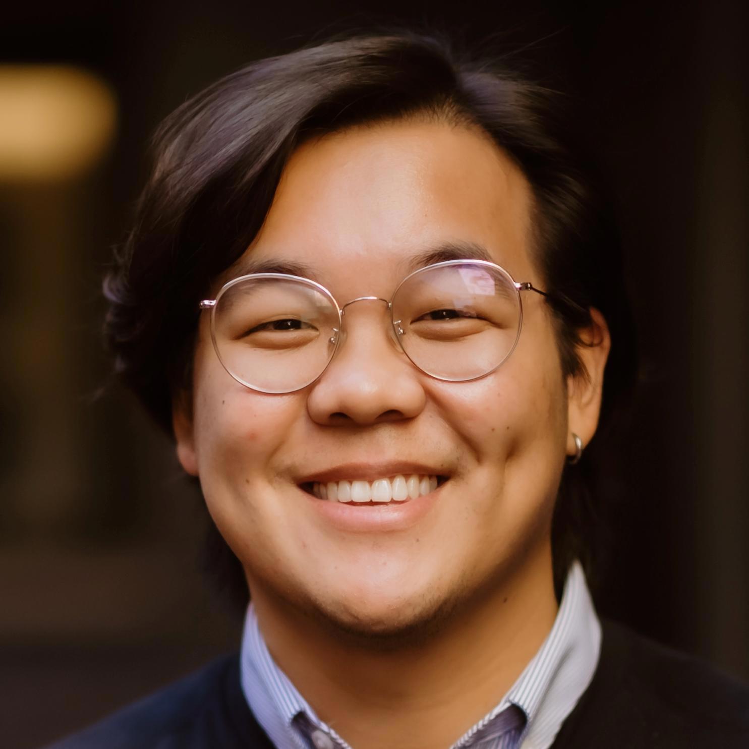 A close-up photo of a young man with medium-length dark hair, wearing round glasses and an earring. He is smiling brightly, showing his dimples. He is dressed in a collared shirt with a dark sweater over it. The background is blurred and dark, drawing focus to his face.