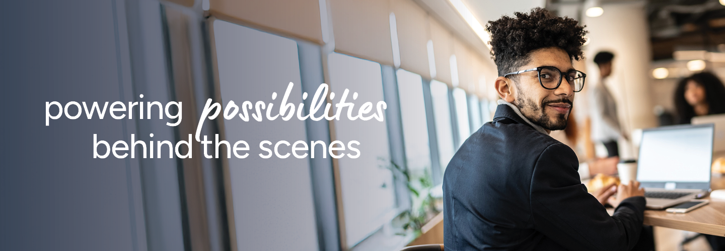 A professional in a suit and glasses sits at a desk with a laptop, smiling and looking over their shoulder. The background shows a modern office setting with colleagues in the distance. The words "powering possibilities behind the scenes" are on the left.