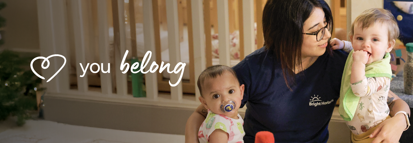 A caregiver in a Bright Horizons shirt is sitting with two infants in a cozy childcare setting. One infant is sucking on a pacifier, while the other is playfully nibbling their hand. The words "you belong" are displayed on the left side.