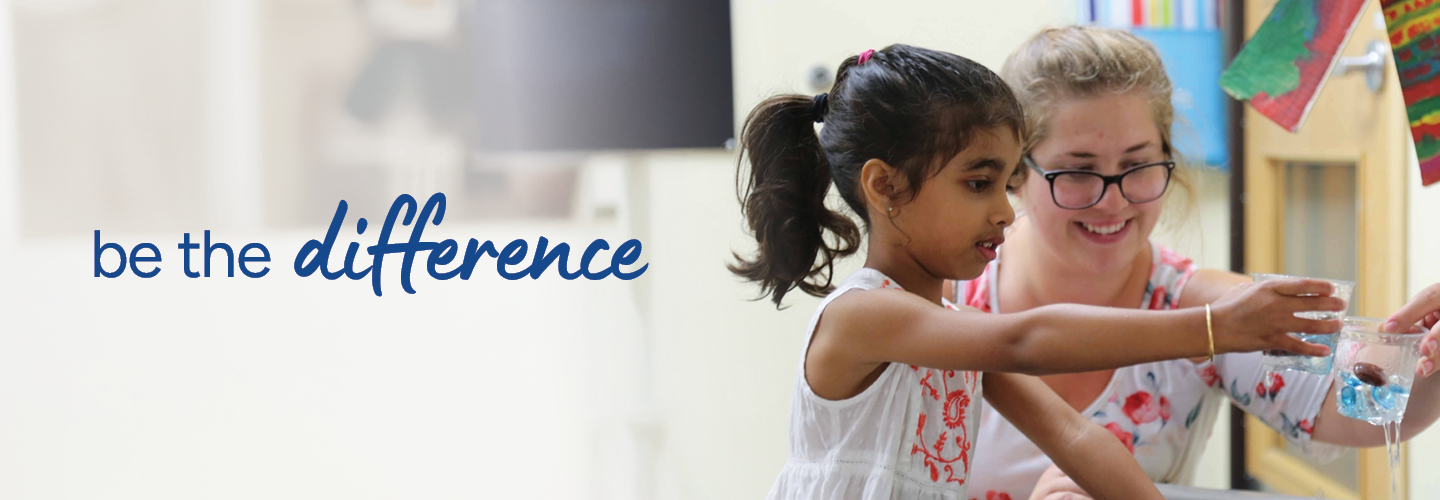 A young girl and a smiling woman engage in a fun water activity at a classroom table. Both are focused on the task, and the girl is pouring water into a cup with floating objects. The words "be the difference" appear on the left.