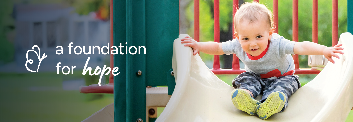 A young child is joyfully sliding down a playground slide. The child is smiling, wearing a gray shirt and camouflage pants. The background is a vibrant playground setting with green grass and colorful structures. The words "a foundation for hope" are on the left side of the image.