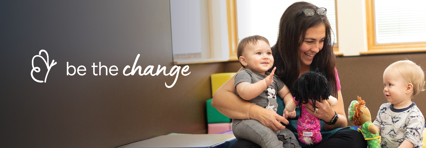 A smiling woman is sitting on the floor, holding two babies in her lap. She is engaging with them using colorful dolls. The background has a soft, inviting playroom setting. The words "be the change" are visible on the left side of the image.