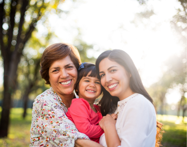 Three generations of women smiling and hugging outdoors in a sunny park setting.