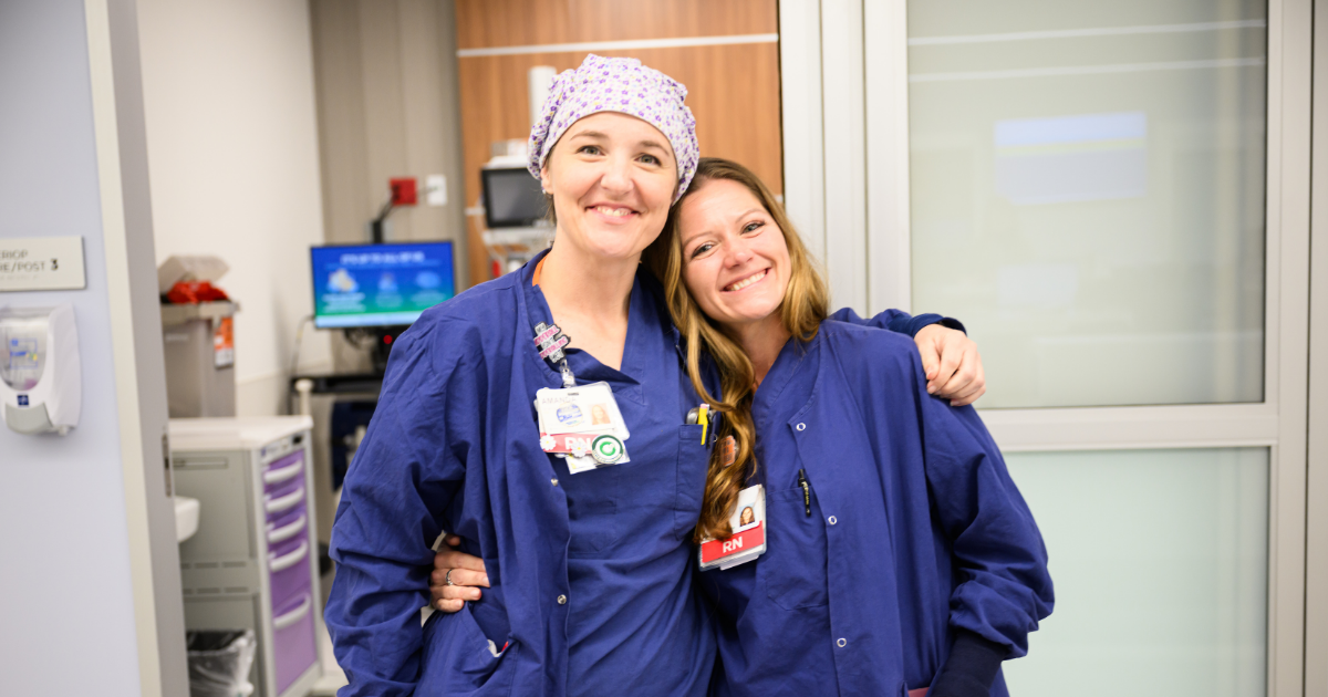 Two smiling nurses wearing navy blue scrubs stand side by side in a hospital setting, with one nurse wearing a patterned surgical cap. They have their arms around each other, showing camaraderie and warmth.