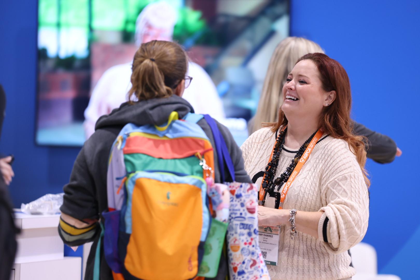 A smiling woman in a beige blouse chats with someone wearing a ponytail and orange backpack.