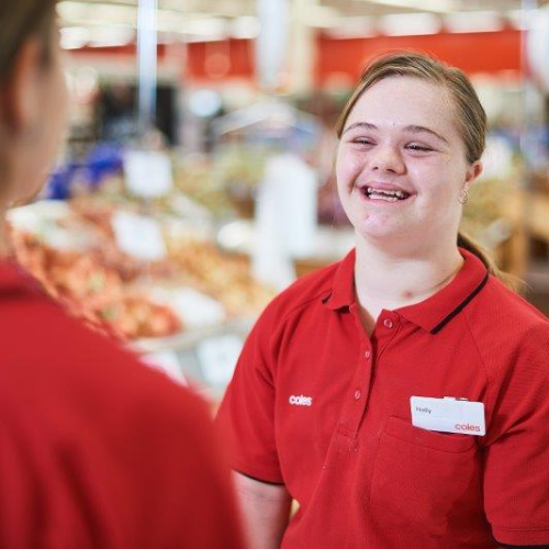A Coles team member with down syndrome in a red uniform with a visible name badge speaking to another person in the checkout section of a Coles supermarket.