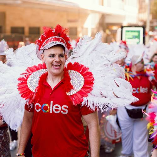 Coles team member wearing a red Coles polo and an elaborate costume with large white and red feathered wings, participating in the Sydney Mardi Gras parade among other colorful participants.