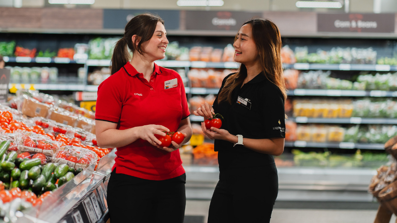 Two female store team members talking to eachother in the fresh produce section of a Coles supermarket. One team member wears a Coles branded red polo while the other wears a black branded Liquorland polo