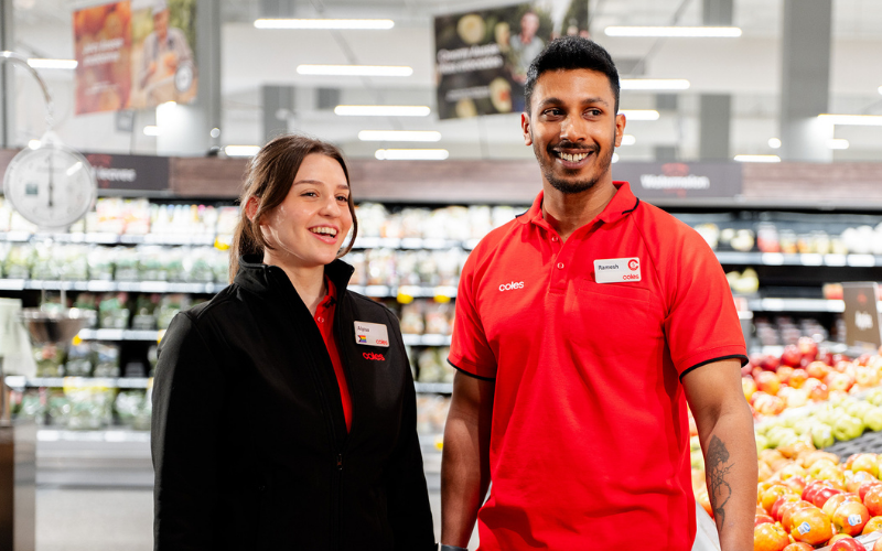 Two Coles team members standing together in the fresh produce section, wearing branded uniforms and name badges, representing teamwork and customer service in a vibrant supermarket environment.