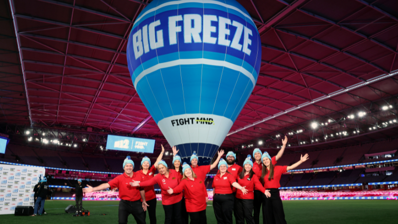 A group of Coles team members in red shirts pose beneath a large Big Freeze hot air balloon on the field at a stadium.