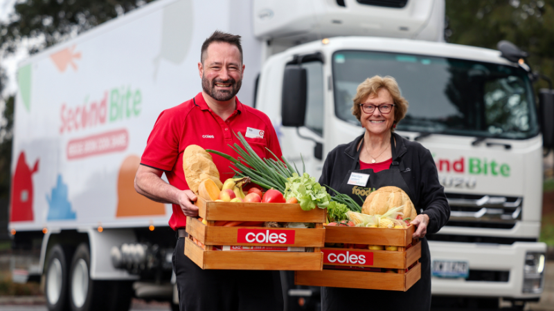 Two smiling people hold wooden Coles crates filled with fresh bread, fruit and vegetables in front of a SecondBite food rescue truck.