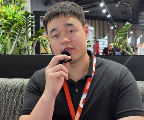 Male in a black polo sits indoors, holding a small clip‑on mic at mouth level; leafy plants and ceiling lights in the background.