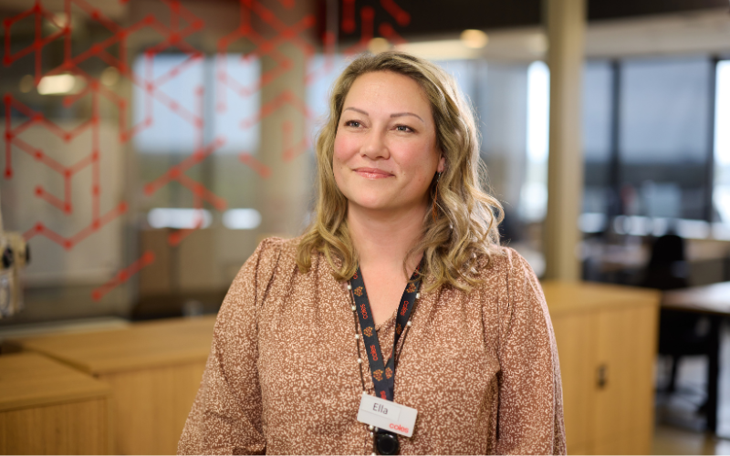 Female wearing a lanyard standing in a modern office workspace with glass walls and desks in the background