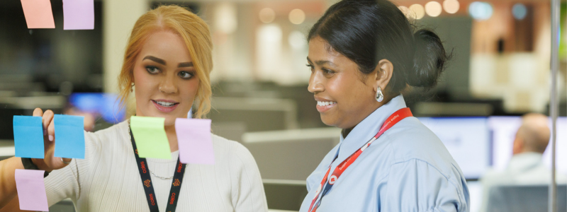 Two colleagues stand in an office workspace, looking at colourful sticky notes placed on a glass surface