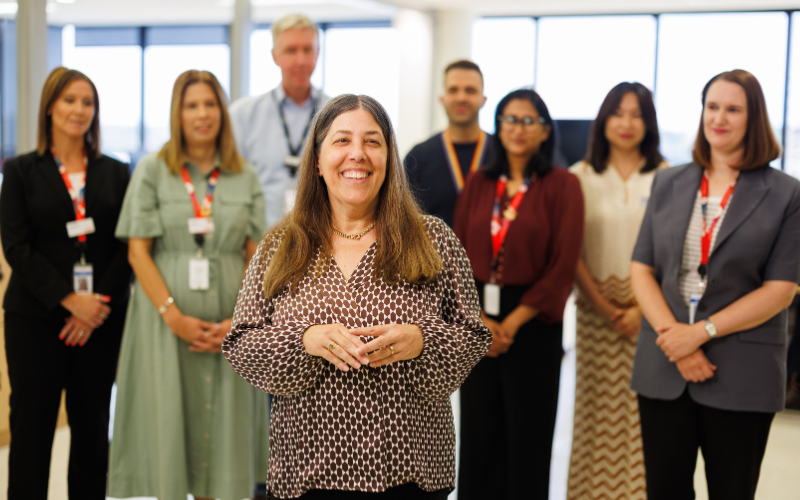 A female stands in the foreground of a bright office space, with a small group of colleagues standing slightly out of focus behind them leading a meeting