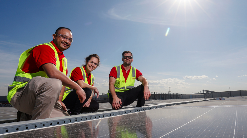Three Coles team members wearing high-visibility vests kneel beside rows of solar panels on a rooftop.