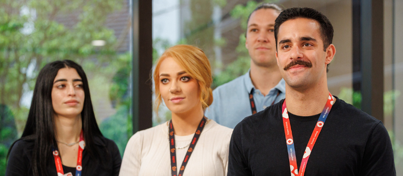 A group of team members standing together in a bright, modern office space with large windows and greenery visible outside. They are wearing company lanyards and name badges, reflecting a professional and collaborative environment.