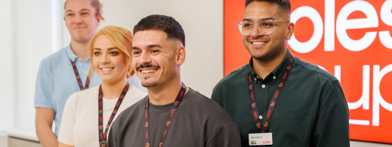 A group of four employees wearing lanyards stand in an office setting, with a red ‘Coles Group’ sign visible in the background