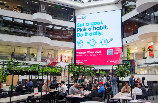 People seated at tables in a large office atrium beneath a digital screen promoting a workplace wellness campaign.