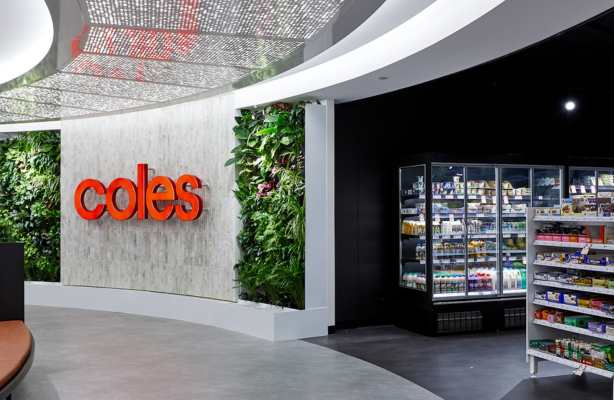 Interior view of a Coles workplace area with a red Coles logo on a wall, indoor green plant features, and a refrigerated food inside the on-site Coles mini store