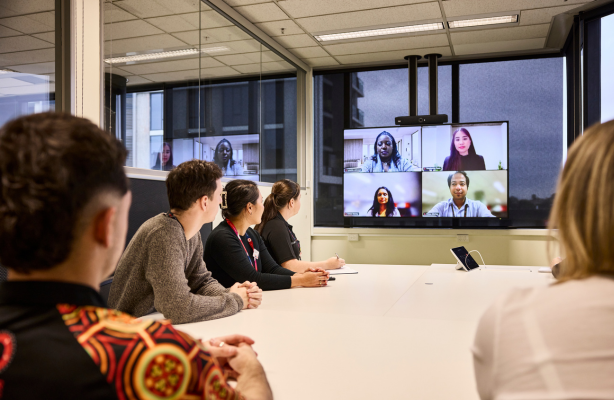 People seated around a meeting table in an office, viewing a video conference with multiple remote participants displayed on a large screen.