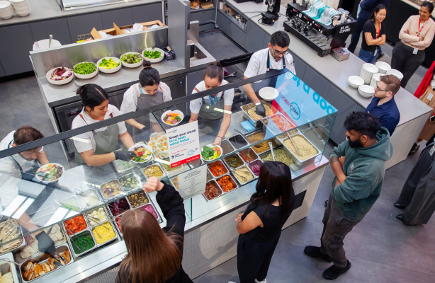 Staff prepare and serve meals behind a glass counter while people select food from trays in a workplace cafeteria.