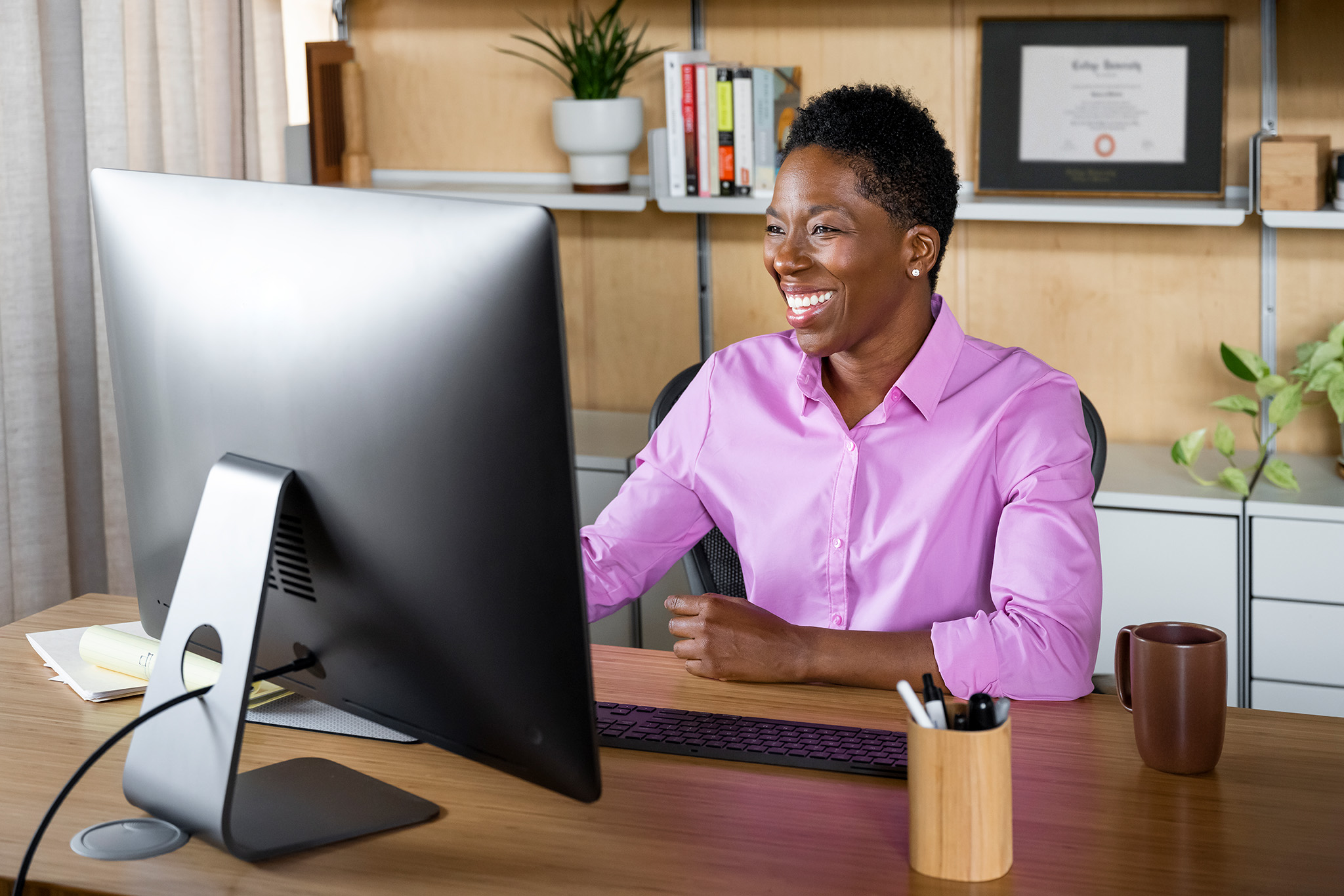 Person in a pink button-down shirt working at a desktop computer in a home office or professional workspace. Books, a framed certificate, and plants are visible on shelves in the background.