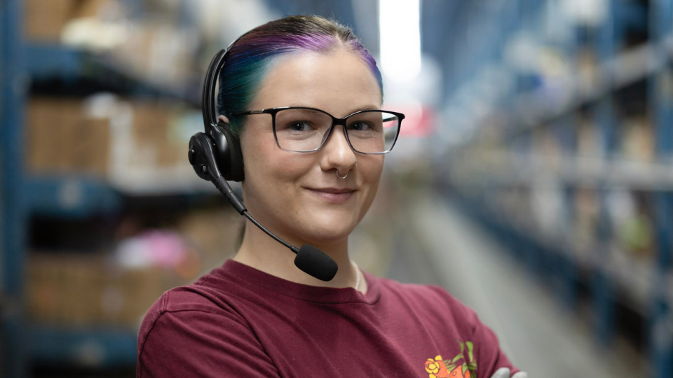 Close-up portrait of Courtney Stockum standing in a warehouse aisle, wearing glasses and a headset with a microphone, arms crossed. The background shows blurred rows of shelving with stored items, emphasizing a busy distribution environment.