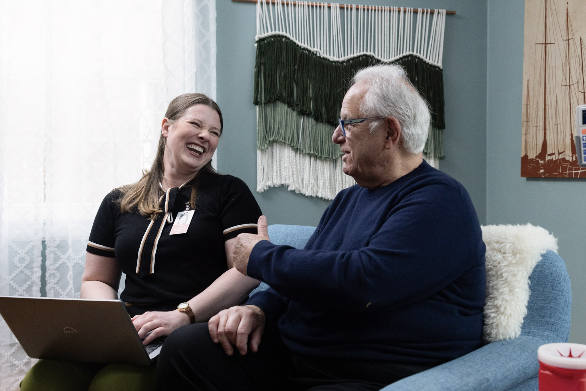 Healthcare professional and patient seated together in a comfortable office setting, engaged in conversation. A blue chair, decorative wall hanging, and framed artwork are visible in the background.