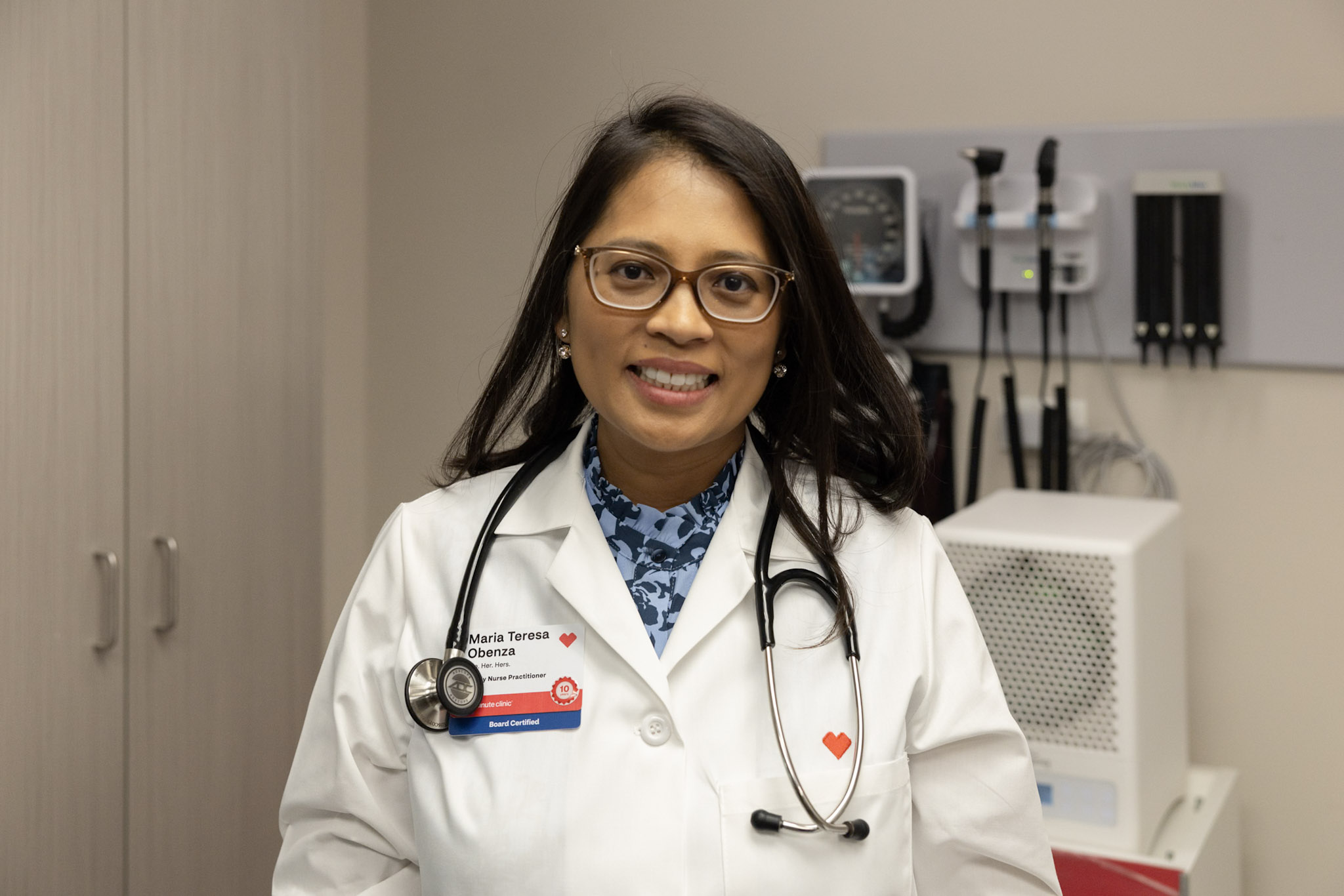 Healthcare provider wearing a white medical coat and stethoscope standing in an exam room. Medical equipment, diagnostic tools, and cabinetry are visible in the background.