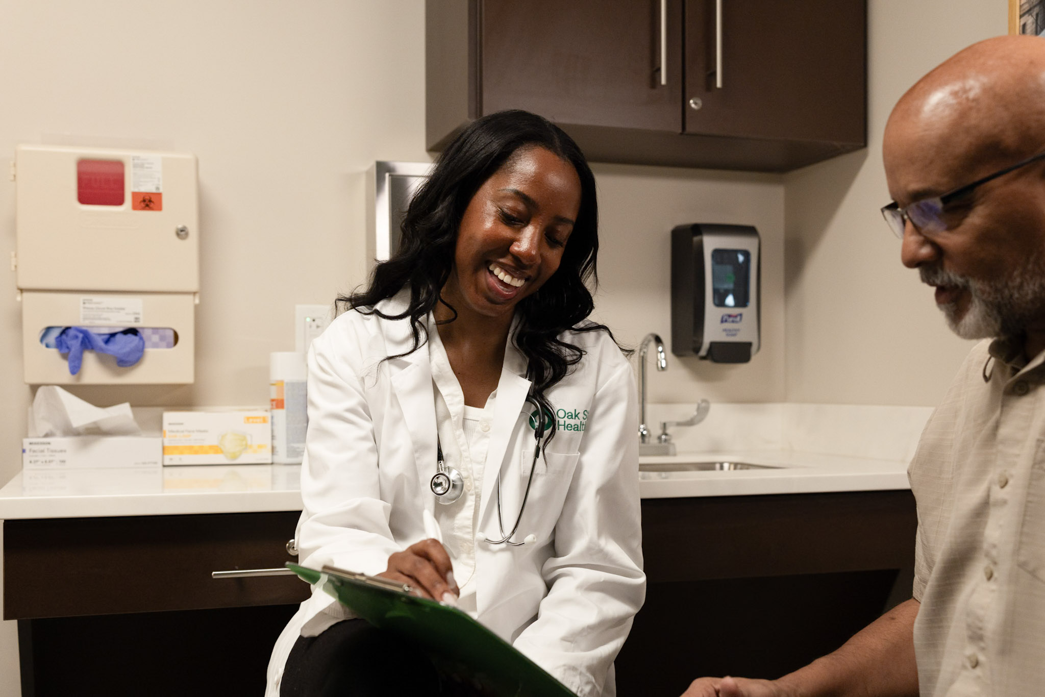 Healthcare provider reviewing paperwork with a patient in a medical exam room. The provider is seated with a clipboard while the patient sits nearby. Cabinets, a sink, medical supplies, and framed wall art are visible in the background.