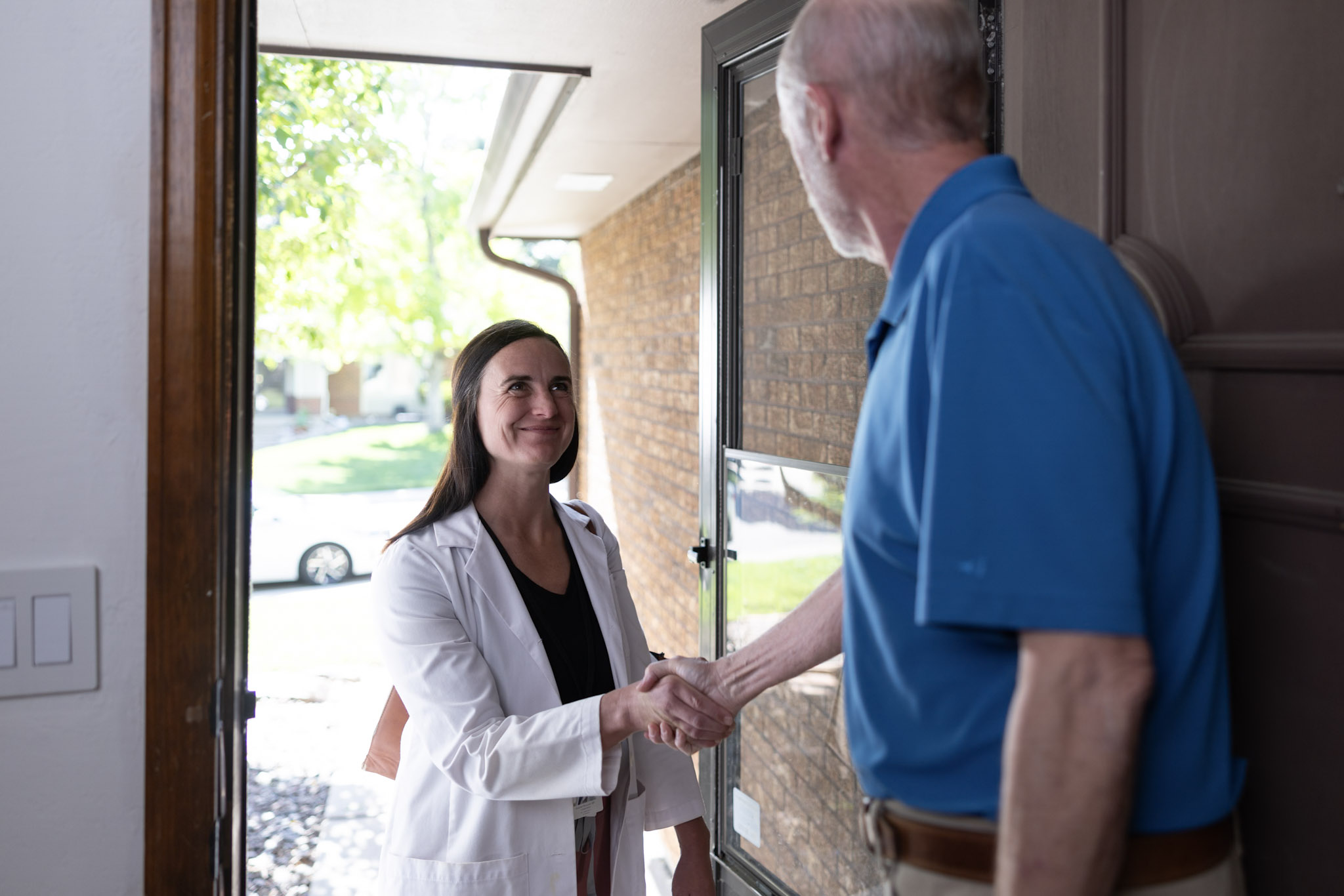 Healthcare professional in a white coat greeting a patient at the front door of a home and shaking hands. The entryway opens to a residential neighborhood, with a brick exterior and natural daylight visible in the background.