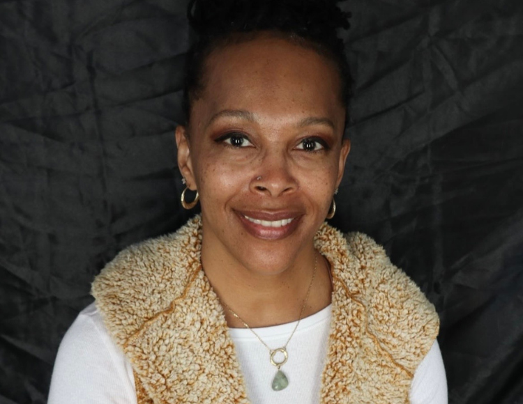 Juanita wearing a textured tan vest over a long sleeve white shirt, sitting in front of a dark, crinkled fabric backdrop. The person has short, dark hair and is wearing hoop earrings and two layered necklaces with small pendants.