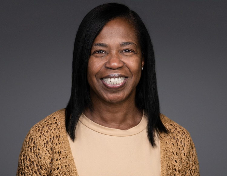 Magnolia wearing a light beige top with a tan, open knit cardigan. The person has straight, shoulder length dark hair and small stud earrings, posed against a smooth, solid gray background.