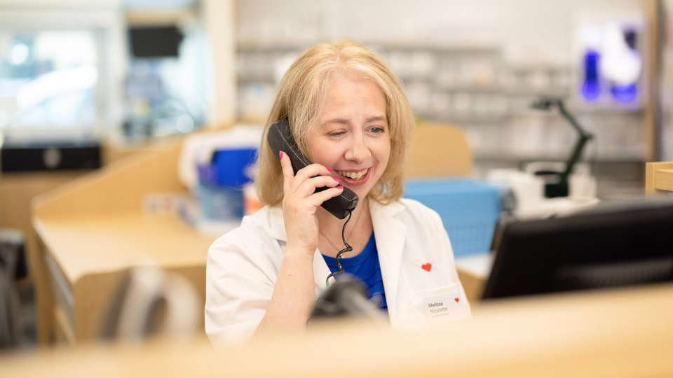 Close-up portrait of Melissa Nicolette wearing a white lab coat, speaking on a corded phone at a pharmacy counter. Blurred shelves, equipment, and workstations in the background suggest a clinical retail setting.