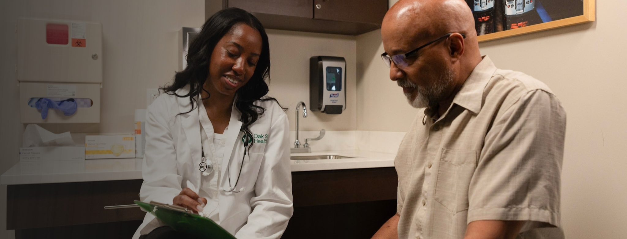Healthcare provider reviewing paperwork with a patient in a medical exam room. The provider is seated with a clipboard while the patient sits nearby. Cabinets, a sink, medical supplies, and framed wall art are visible in the background.