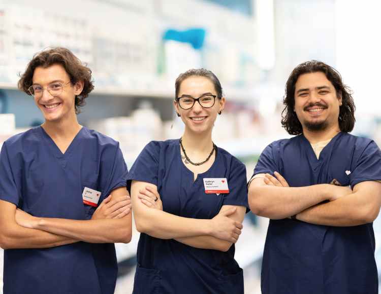 Three CVS Health colleagues wearing navy scrubs stand side by side with arms crossed in a pharmacy setting. The background shows blurred shelves and supplies, suggesting a clinical or retail healthcare environment.