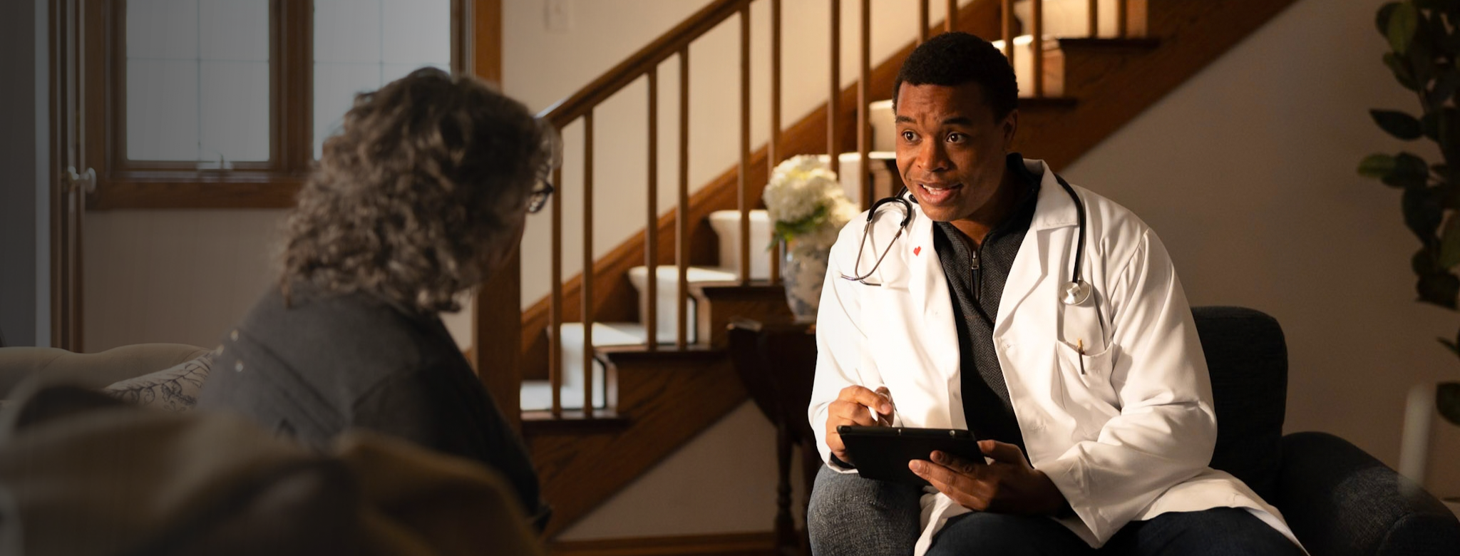 Healthcare provider in a white medical coat and stethoscope conducting an in-home visit with a patient. The provider is seated with a tablet in a residential living room, with a staircase and home furnishings visible in the background.