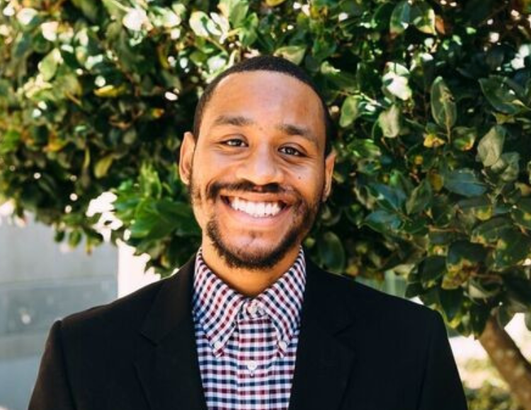 Quenton wearing a checked button up shirt and a dark blazer, standing outdoors in front of a leafy green tree. Part of a building structure is visible in the background.