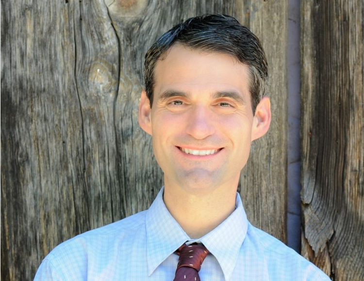 Tony wearing a light blue dress shirt and a patterned necktie, standing in front of a weathered wooden wall with visible grain and knots.